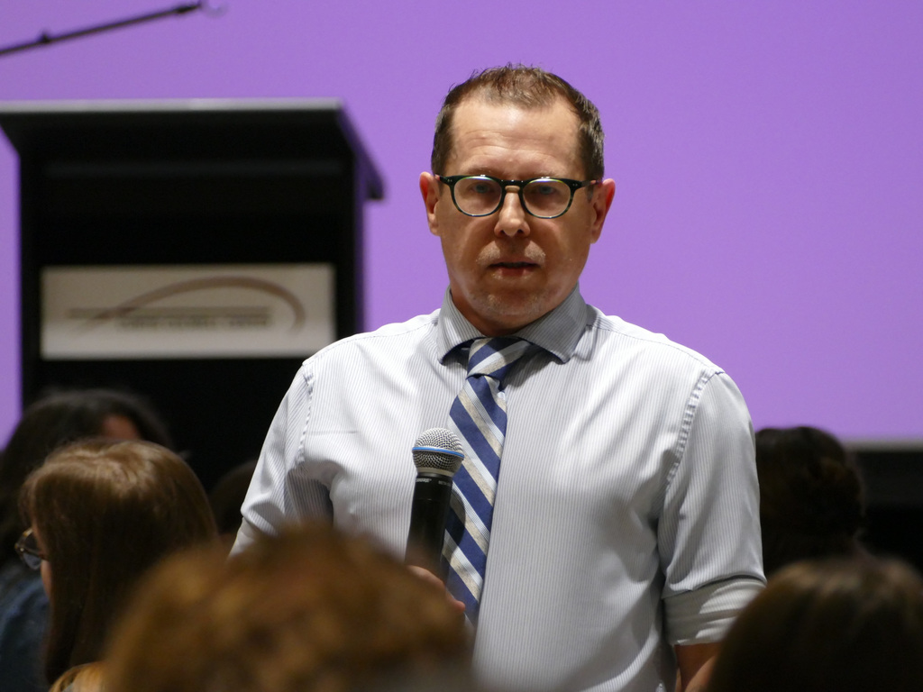 Person speaking at a podium with a microphone in a room with a purple background.