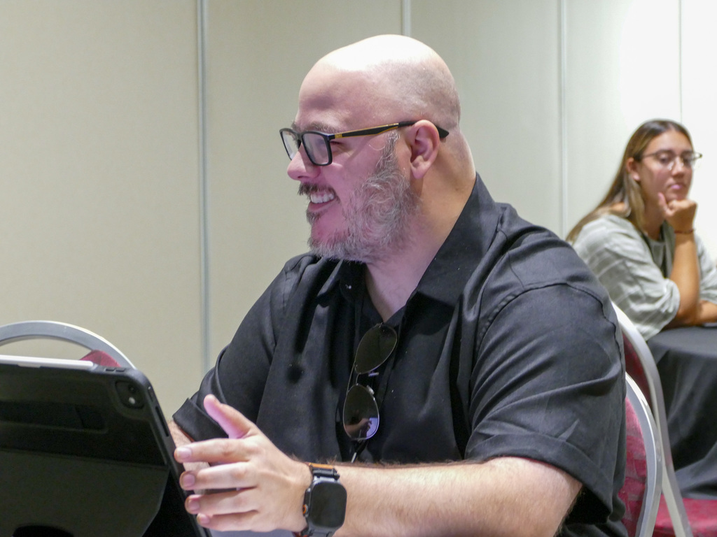 Person at a conference holding a smartphone and smiling, seated in a meeting room.