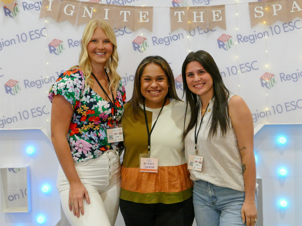 Three individuals standing and smiling at an event in front of a banner that reads "Ignite the Spark Region 10 ESC." All are wearing name tags and dressed in smart casual attire.