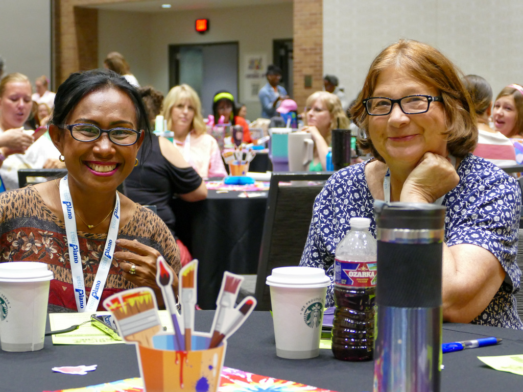 Two ladies smiling at the camera, sitting in a conference, both are wearing colorful shirt and glasses.