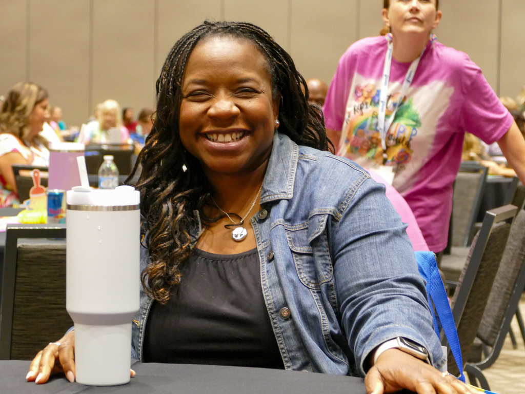 A lady wearing a jeans jacket and s black shirt sitting at a conference while smiling at the camera