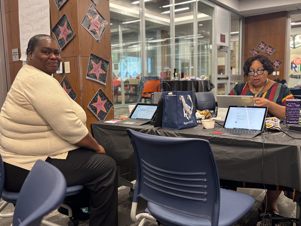 Two individuals are seated at a desk in a library, each working on a laptop. There are snacks and personal items on the desk, and educational posters are visible on the glass wall behind them.