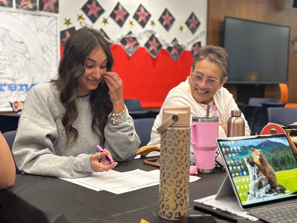 Two people smiling and taking notes at a table during a meeting, with a laptop showing a nature image in the foreground. There are personal items like water bottles and a bag visible, set in a room decorated with a wall emblem that reads "Parents.
