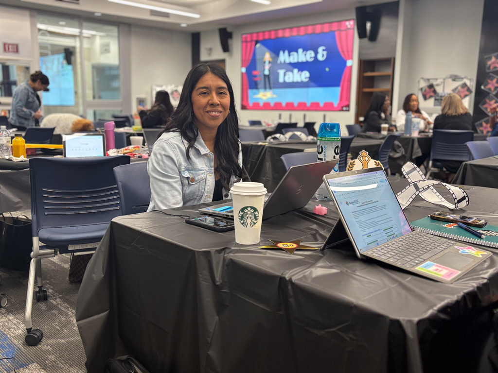 Person sitting at a table with a laptop, coffee cup, and crafting supplies, in a conference room during a "Make & Take" event.