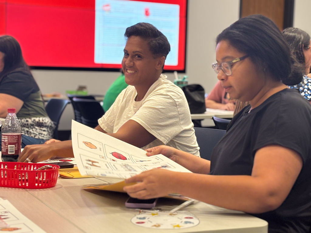 An educational workshop at a university, featuring participants seated at tables reading printed materials, with a person smiling at the camera.