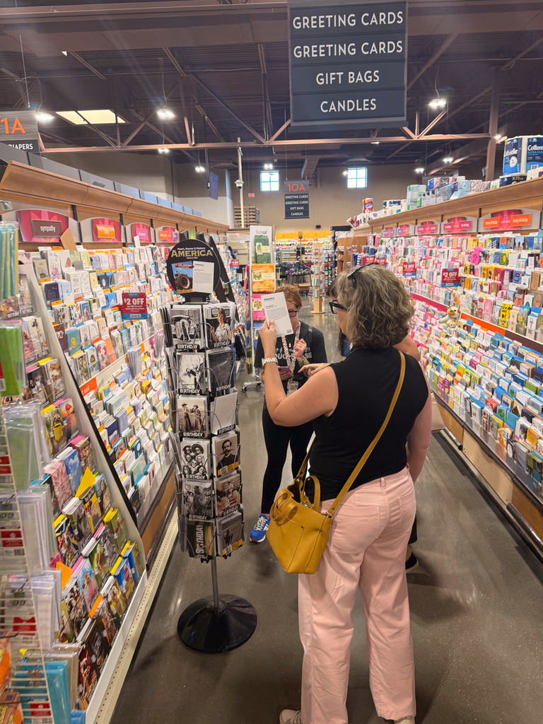 Person browsing greeting cards in a supermarket aisle, with another person in the background also looking at cards.