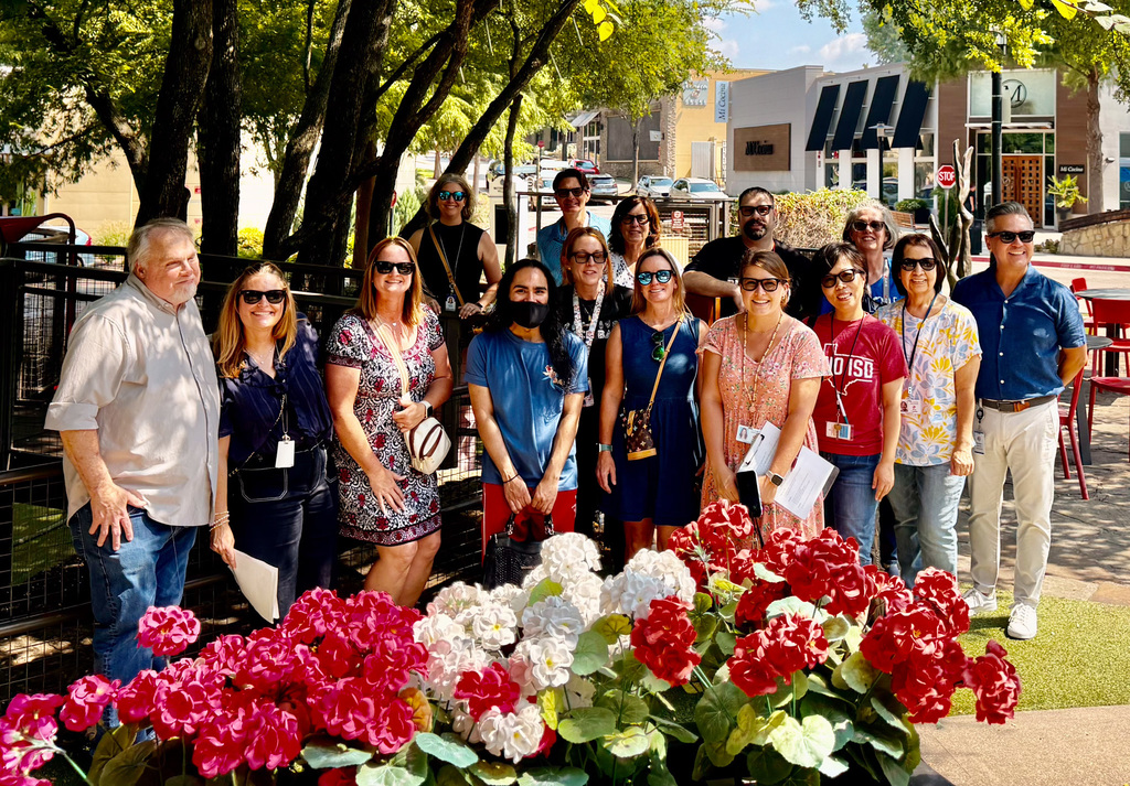 Group of people posing for a photo outdoors, in front of vibrant red and white flowers, with a street and buildings in the background.
