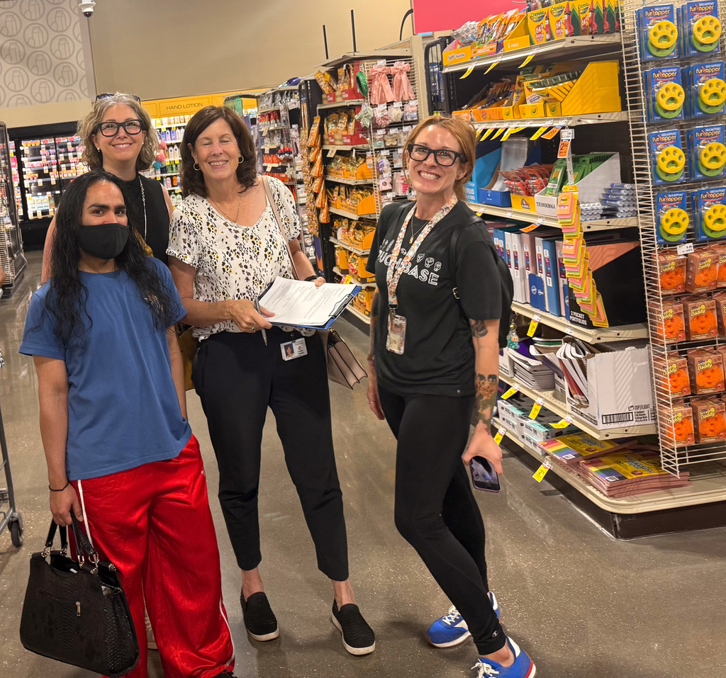 Four people standing in a grocery store aisle, smiling at the camera, surrounded by shelves stocked with various products.