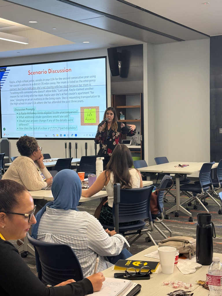 A presenter discussing a scenario at a workshop in a conference room, with attendees seated at tables focusing on the presentation. The screen behind the speaker displays text and bullet points.