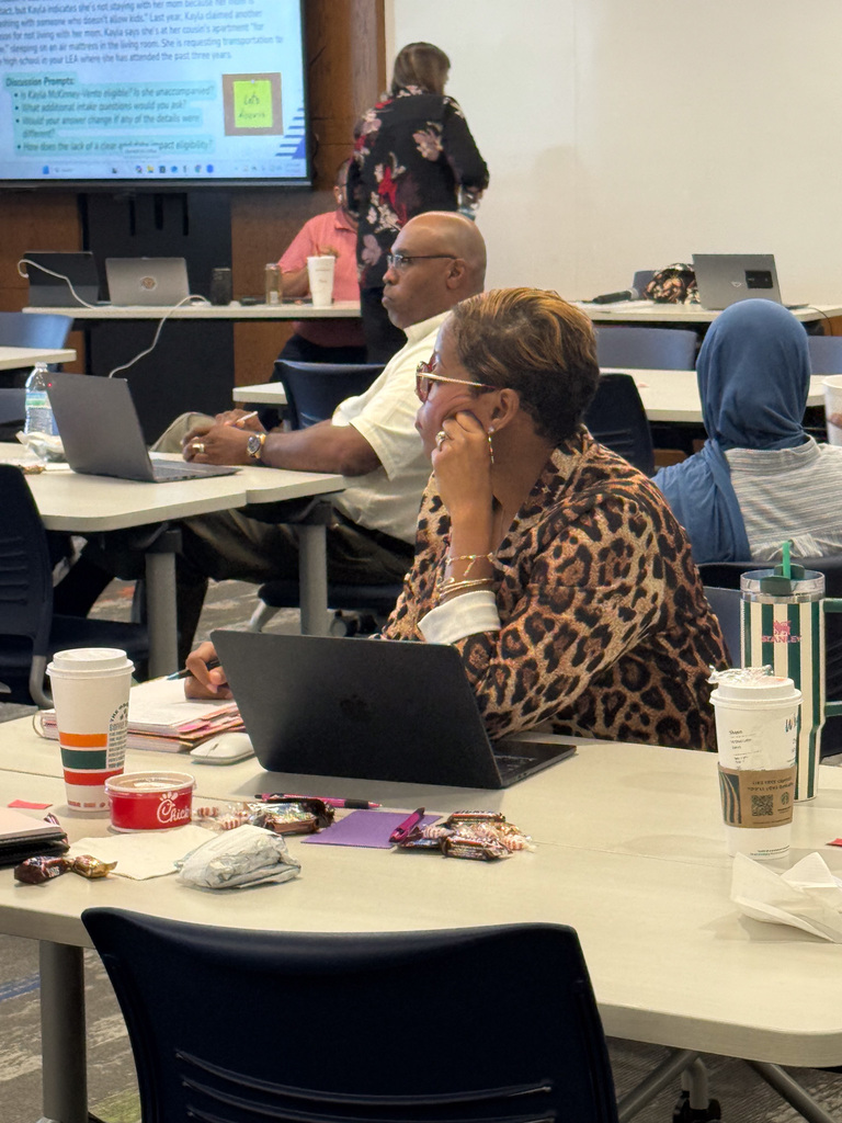 Participants attending a workshop, seated at tables with laptops and personal items, focused on a presentation in a room with a projector screen displaying text.