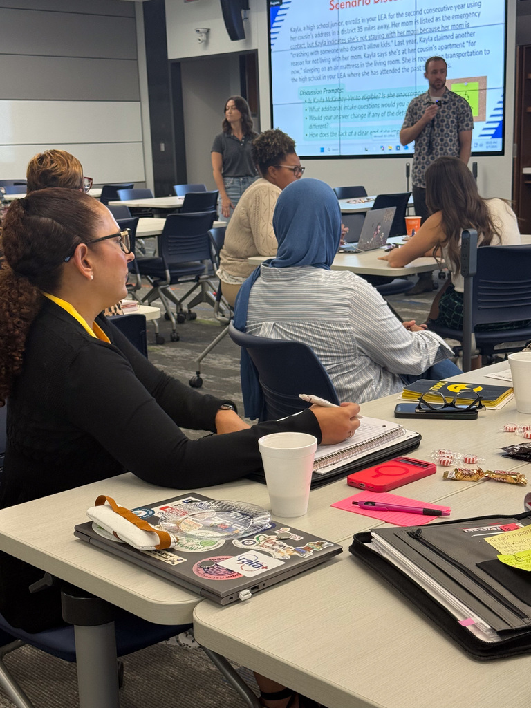 A group of professionals at a workshop, with one person leading a presentation at the front. The room has modern furnishings and several attendees using laptops.
