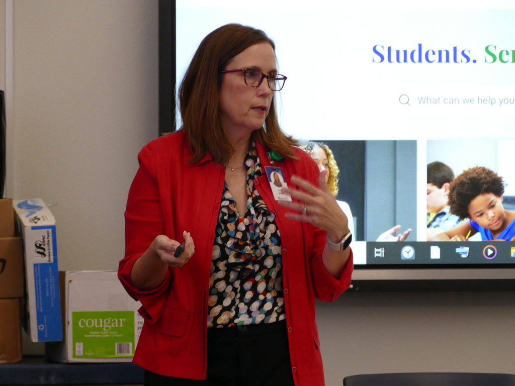 A person giving a presentation in front of a screen displaying the word "Student." The presenter is wearing glasses and a red blazer over a patterned blouse. There is a name badge visible on the blazer.