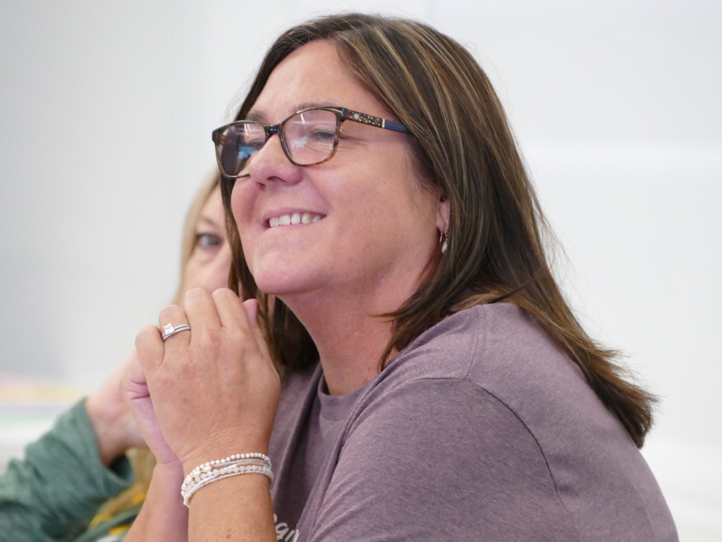 Close-up of a smiling person wearing glasses and a purple shirt, with hands clasped near the chin.