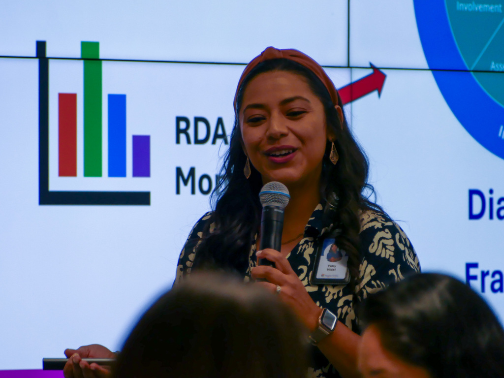 A person giving a presentation using a microphone, standing in front of a screen displaying various graphs and the RDA Monthly logo.