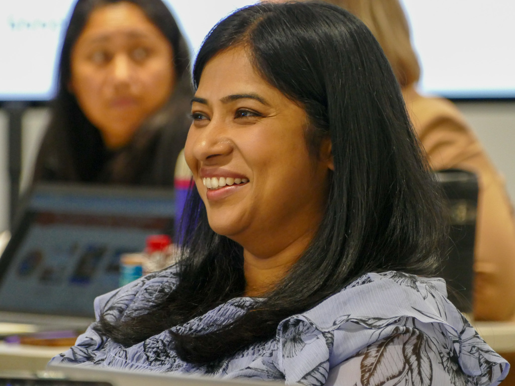 An individual smiling during a meeting with other attendees in the background.
