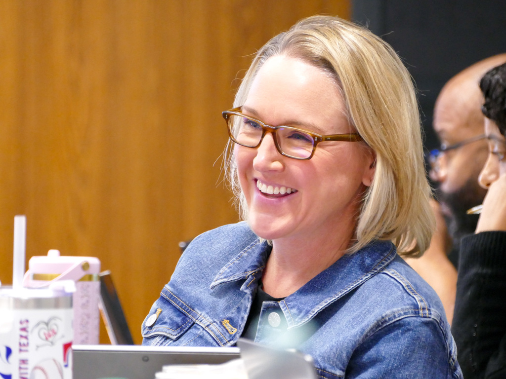A person wearing glasses and a denim jacket is smiling and looking away from the camera, sitting at a table with a soda cup and laptop, in a room with wood paneling and another person visible in the background.