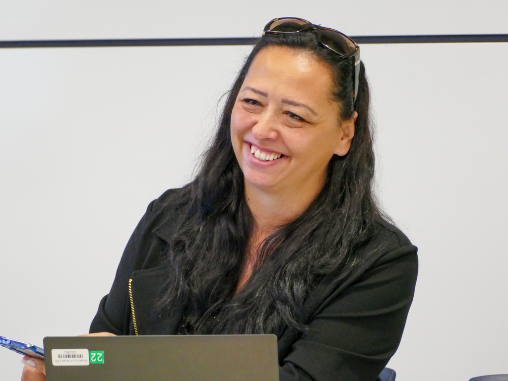 Person smiling and sitting at a table with a laptop, participating in a meeting. There is a notebook and a pen also visible on the table.