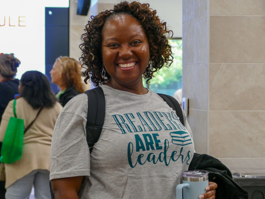 A person wearing a t-shirt with the text "Readers are Leaders" smiles at the camera in a busy indoor location.