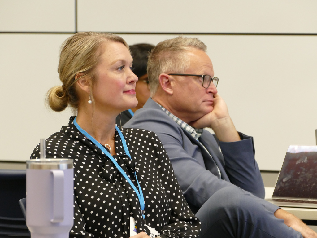 Two attendees listening intently at a conference. One is wearing a polka dot shirt and a lanyard.