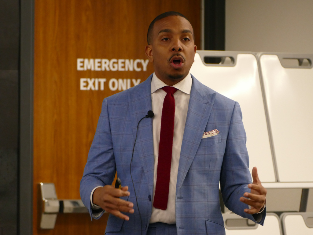 Person giving a presentation in a conference room with a sign saying "EMERGENCY EXIT ONLY" in the background. The individual is dressed in a blue suit with a red tie and is gesturing with their hands.