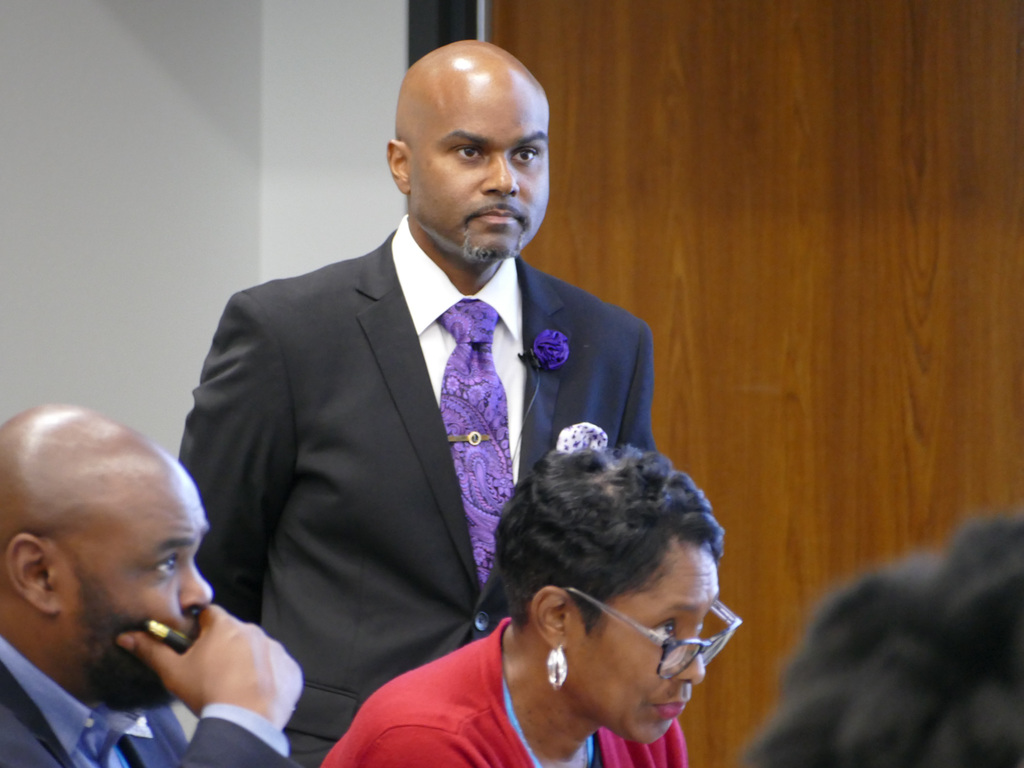 Person standing and listening attentively during a meeting, wearing a formal suit with a purple tie and flower pin.