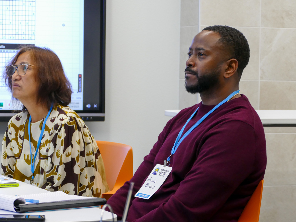 Two attendees, one wearing a name tag labeled "Anisa" and the other labeled "Hans", are seated at a conference, listening attentively.