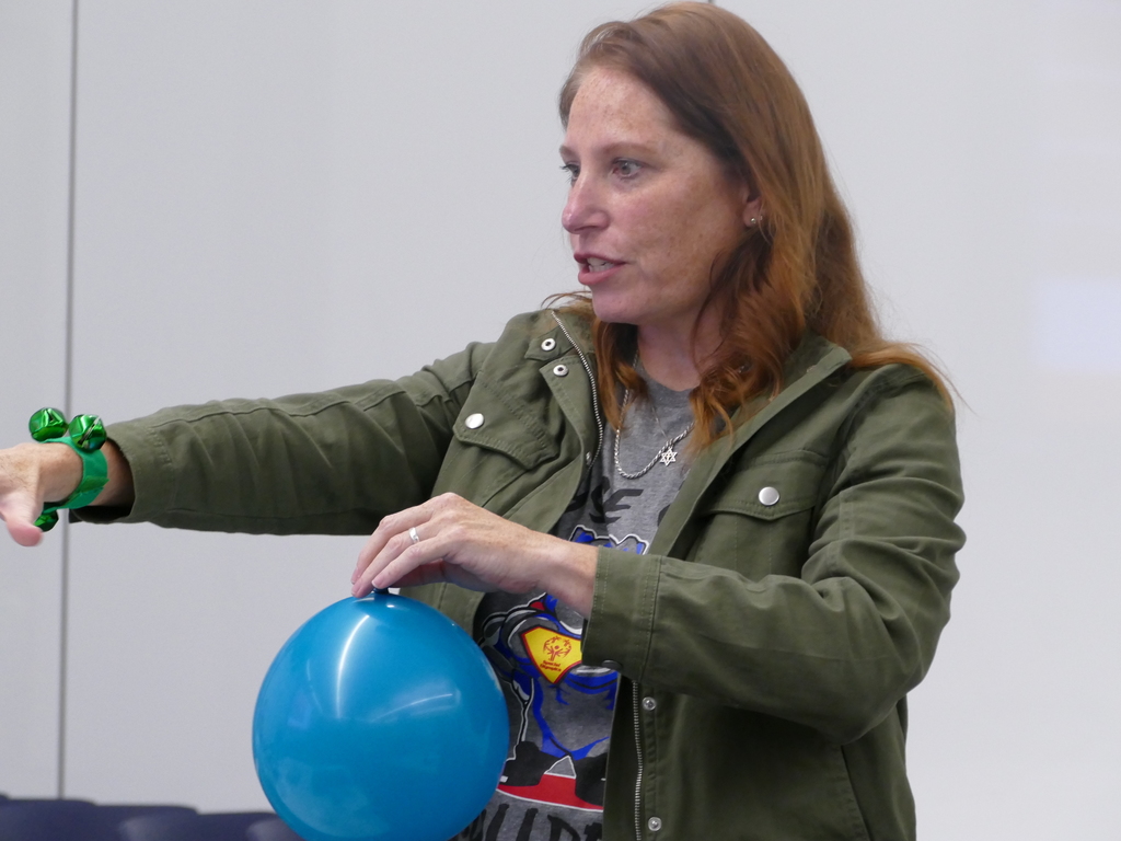 A person wearing a Superman T-shirt and a green jacket is demonstrating with a blue balloon in a classroom setting.