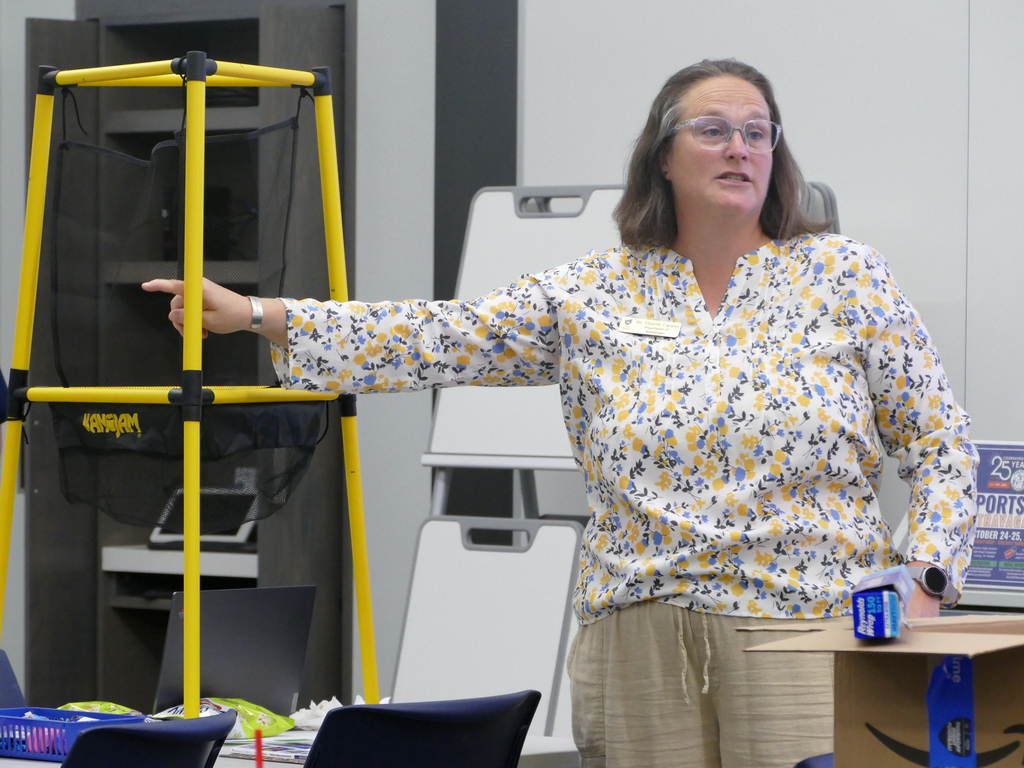 A person is giving a presentation in a classroom, standing next to a digital screen and pointing at a flip chart. There are books and a blue can on a table nearby.