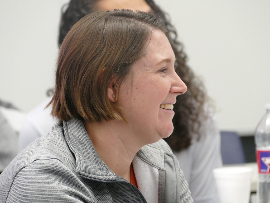 Person smiling and sitting in a meeting room, with a partial view of another attendee and a water bottle labeled "Pepsi."