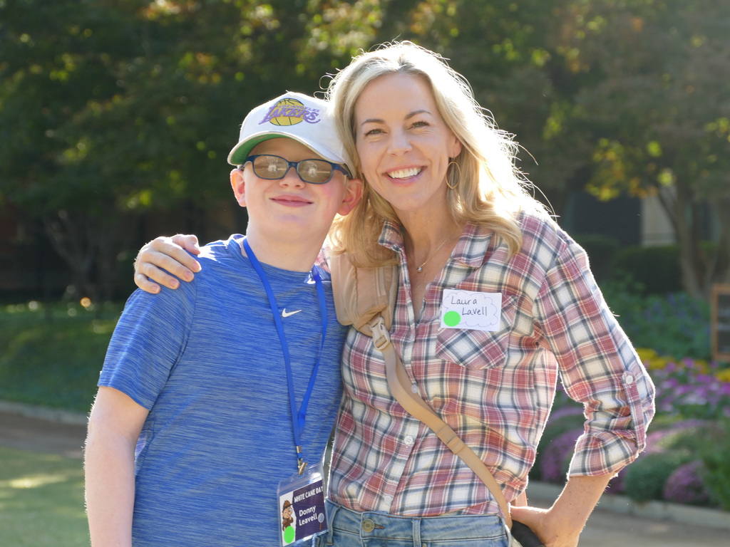 Two smiling individuals, one wearing a blue shirt and a hat labeled "LSU," and the other in a plaid shirt with a name tag reading "Laura," embracing outdoors on a sunny day.