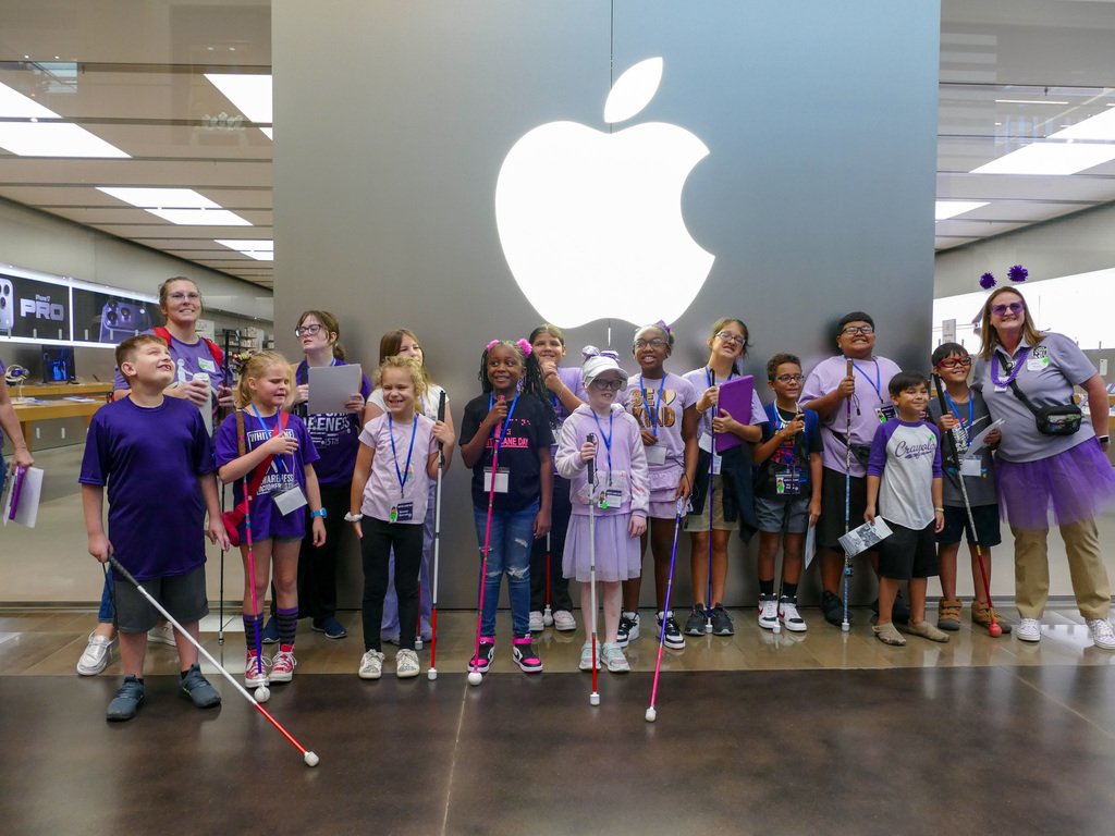 Group of children with a couple of adults posing in an Apple Store, standing in front of a large lit Apple logo, some wearing purple shirts and lanyards.