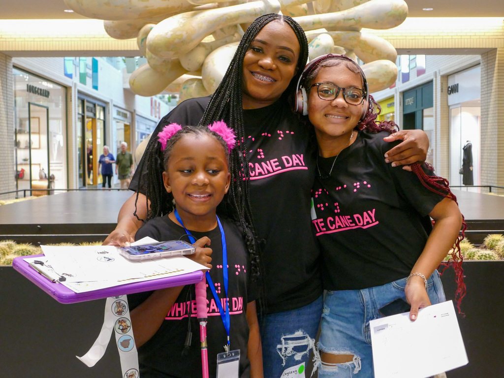 Three people wearing matching "White Cane Day" T-shirts smiling together in a shopping mall.