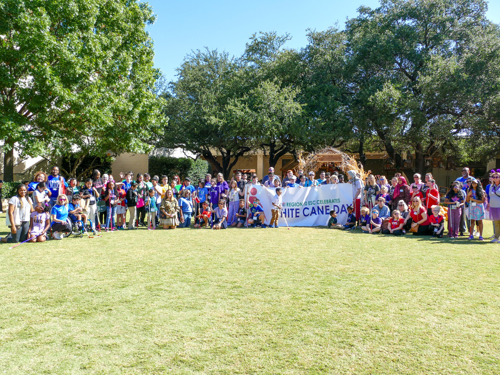 Group of people gathered outdoors at a White Cane Day event, holding banners and signs, with trees in the background.