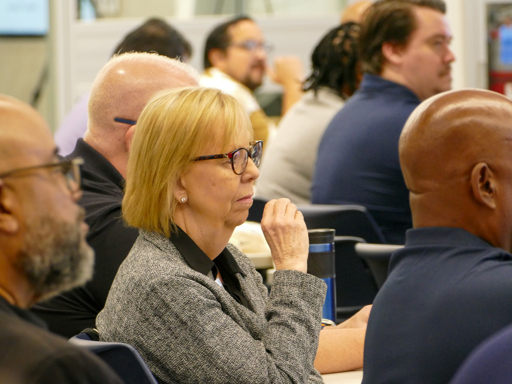 A lady as the audience of a presentation, listening attentively, she's wearing glasses and a gray jacket. 