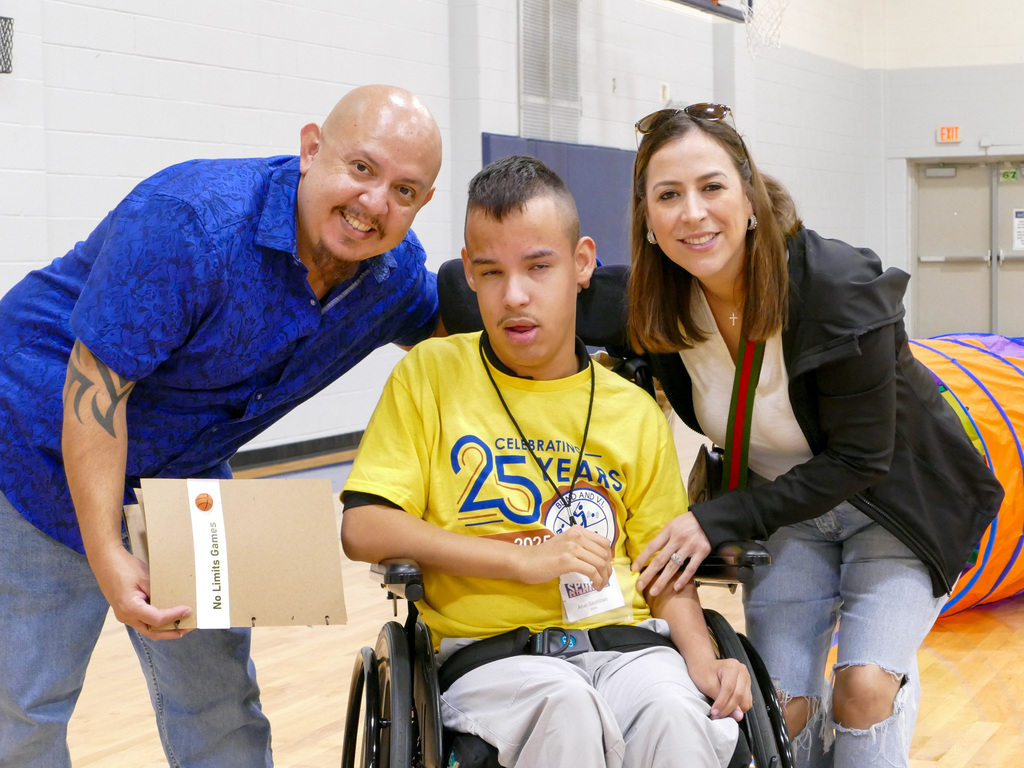 Two adults smiling and standing beside a seated individual in a wheelchair at a Special Olympics event, all are holding diplomas.