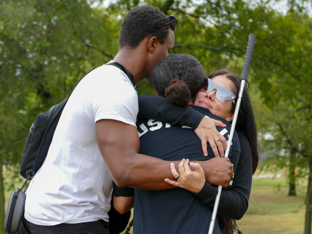 Three individuals share a heartfelt embrace in a park, one of them holding a white mobility cane.