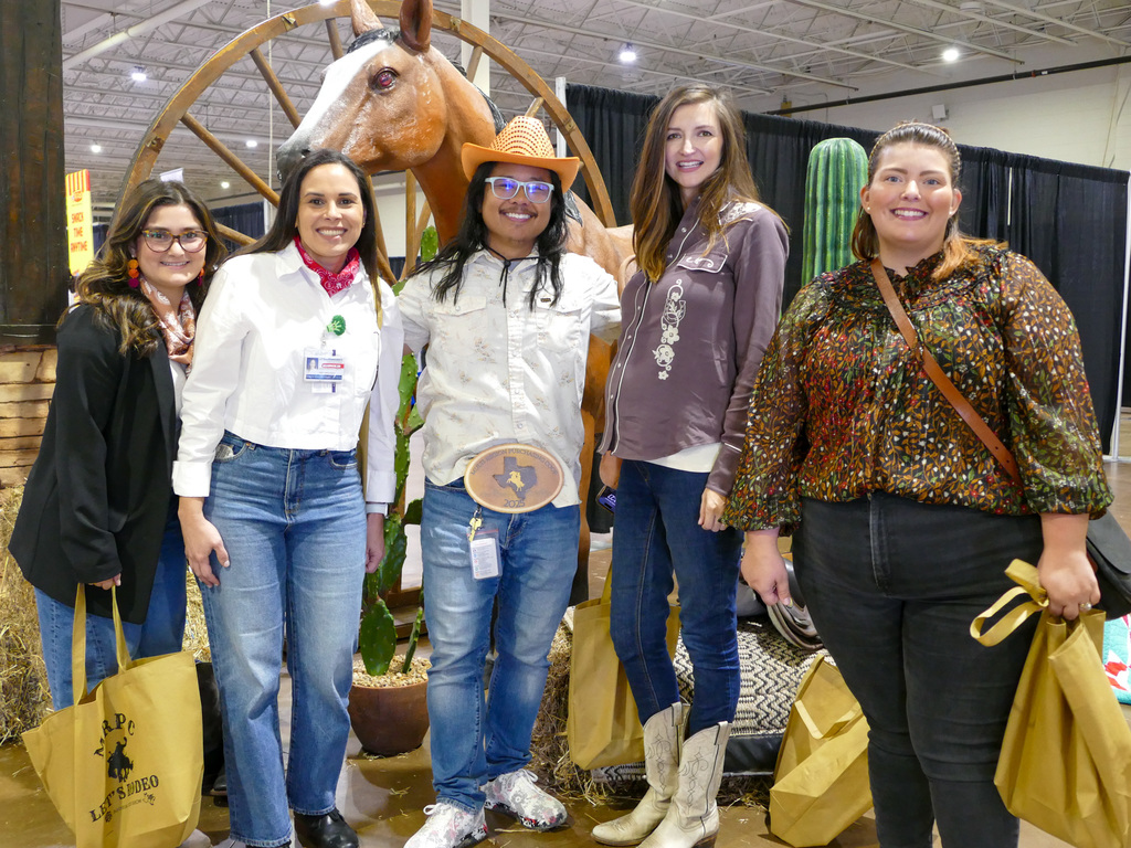 Five attendees at a Texas-themed event, standing in front of a large wooden wheel and a model horse. Each person is dressed in different styles, including hats and jeans, and holding event bags.