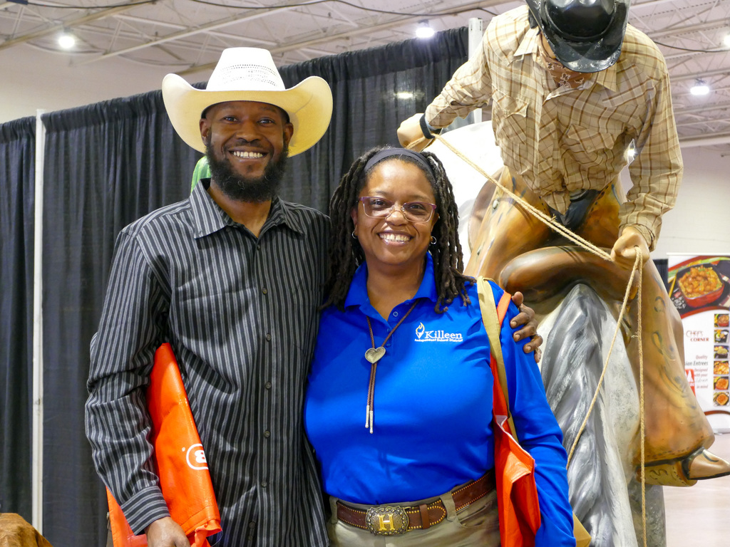 Two individuals posing with a smile at an exhibition booth, one wearing a cowboy hat and the other dressed in a blue 'VisitLEX' polo shirt, standing next to a mannequin on a horse.