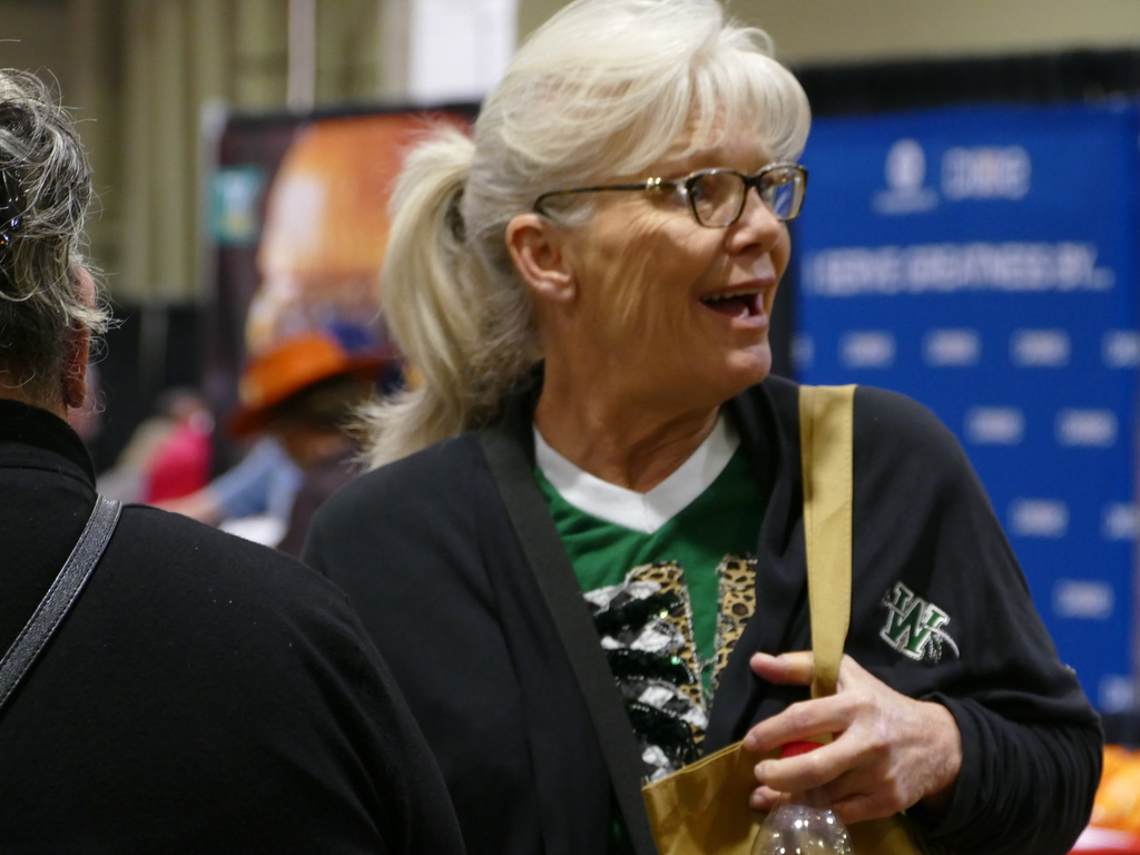 Two individuals are engaged in a conversation at a convention, with one person wearing a festive sweater and glasses, and the backdrop features a banner with the words "Duke Energy."