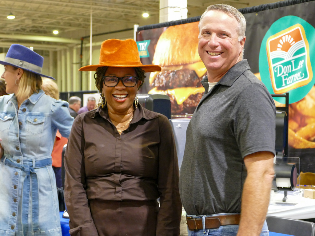 Two smiling people at a trade show booth with a "Don Lee Farms" banner in the background. One person is wearing a stylish orange hat, and the other is in a casual blue shirt.