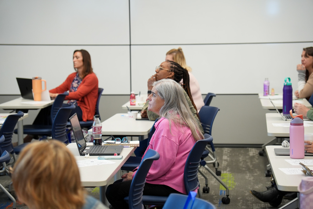 Adults seated at tables in a classroom setting, engaging with laptops and conversing with each other.