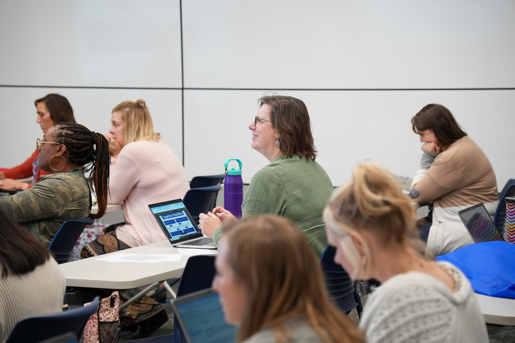 Adults seated at tables in a classroom setting, engaging with laptops and conversing with each other.