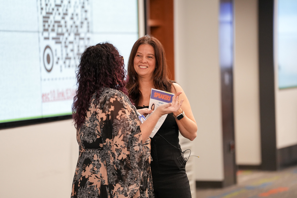 Two individuals engaging in a conversation at a conference, with one holding a 'SPEAKER' badge. A QR code is displayed in the background on a large screen.