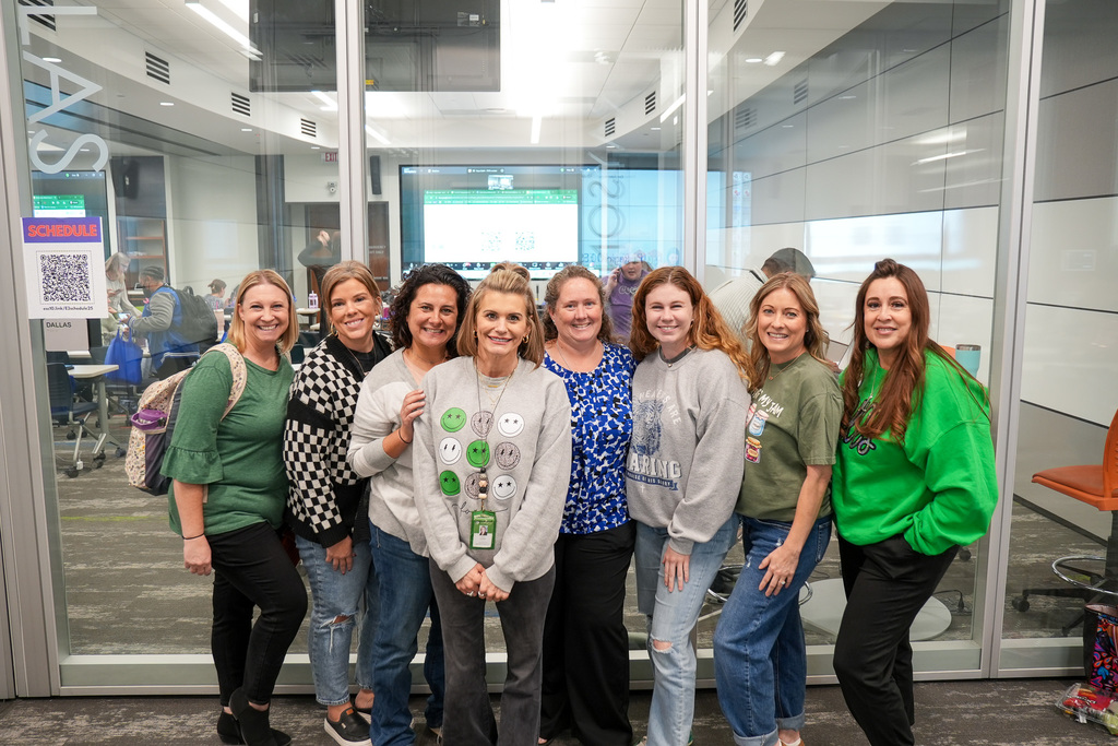 Group of eight individuals posing in an office environment, smiling, with a glass door in the background showing the interior of a meeting room. There are visible signs and laptops