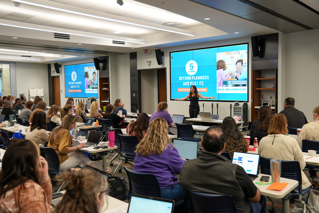 A professional development seminar in progress with attendees seated at tables facing a presenter near a screen displaying a slide titled "BEYOND PLANKS AND ASSISTS".