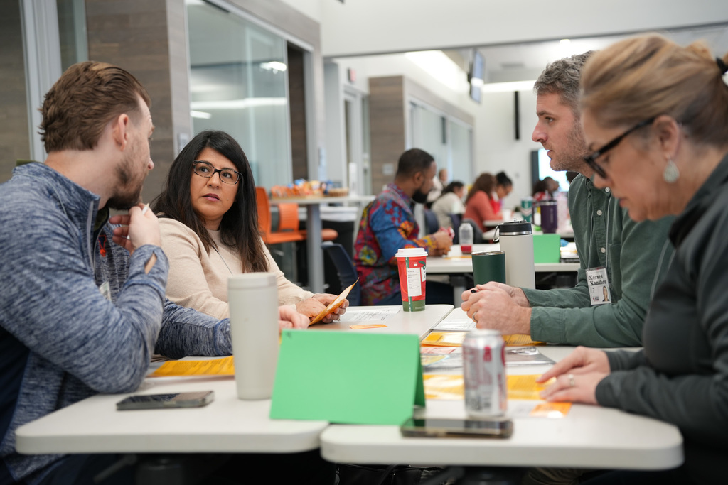 Four adults are seated around a table in a brightly lit cafeteria, engaged in a discussion with notebooks and papers in front of them.