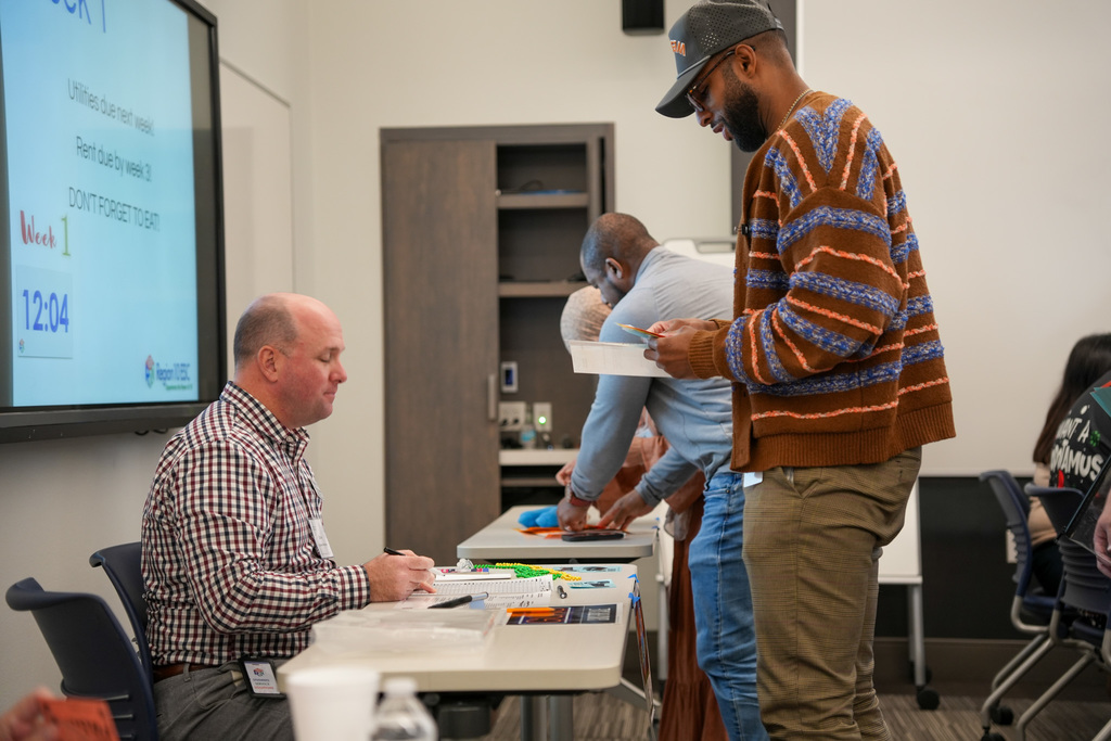 Two individuals discussing a document at a table during a workshop, while another person interacts with a participant in the background. A presentation screen shows a timetable titled "DONTFORGET TO USE.