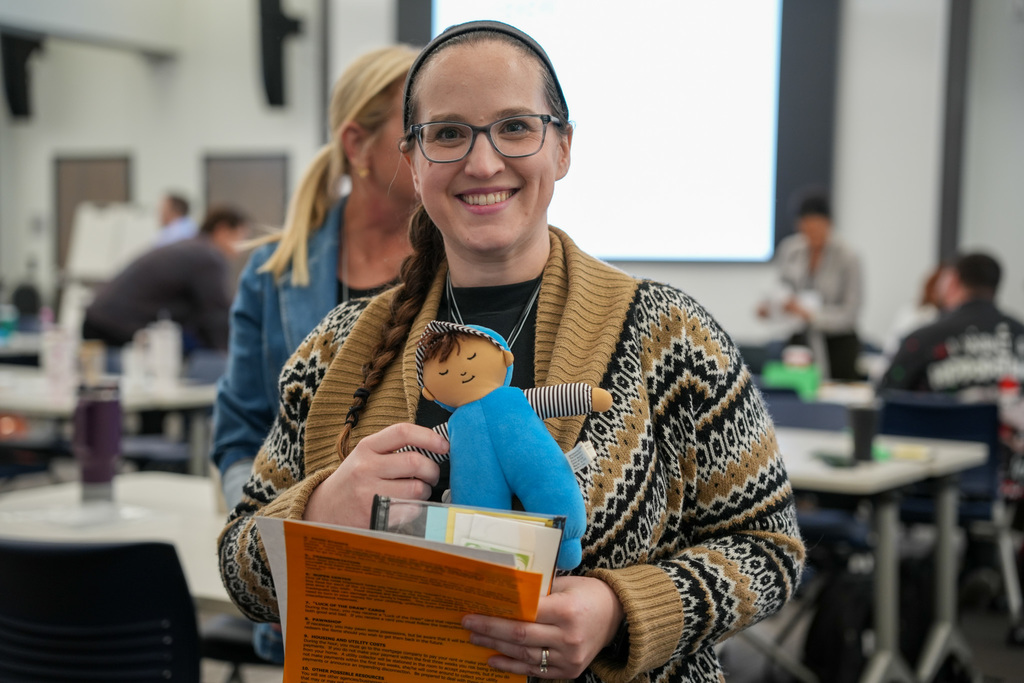 Person smiling and holding a blue doll at a workshop, with attendees and a projector screen in the background.