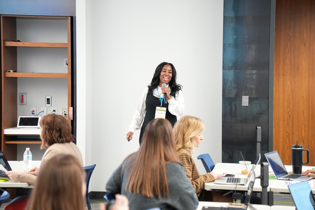 A person presenting to a group of attentive listeners in a modern conference room setting. The speaker holds a microphone and smiles broadly, standing in front of a projected screen and beside a bookshelf. Audience members, seated at tables equipped with laptops, focus on the presentation.