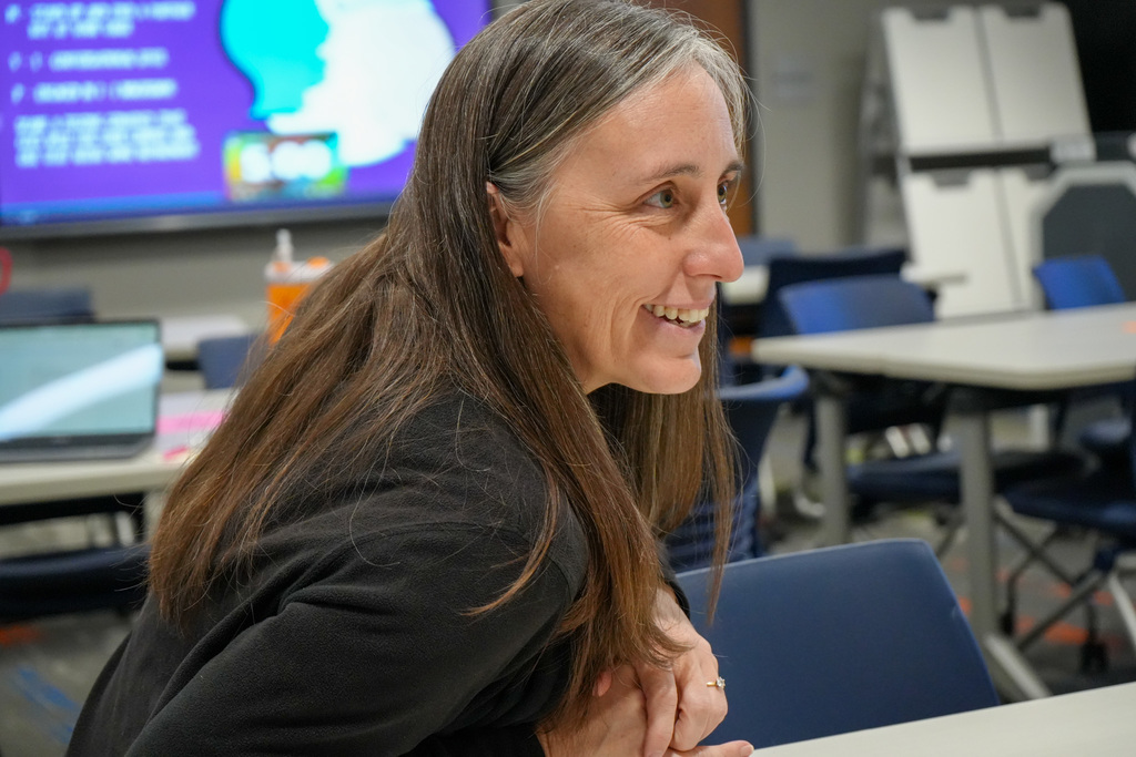 Person smiling in a classroom with a digital screen displaying graphics in the background.